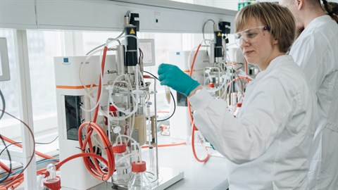 En kvinde i hvid kittel og blå gummihandsker arbejder ved en lille bioreaktor i et laboratorie