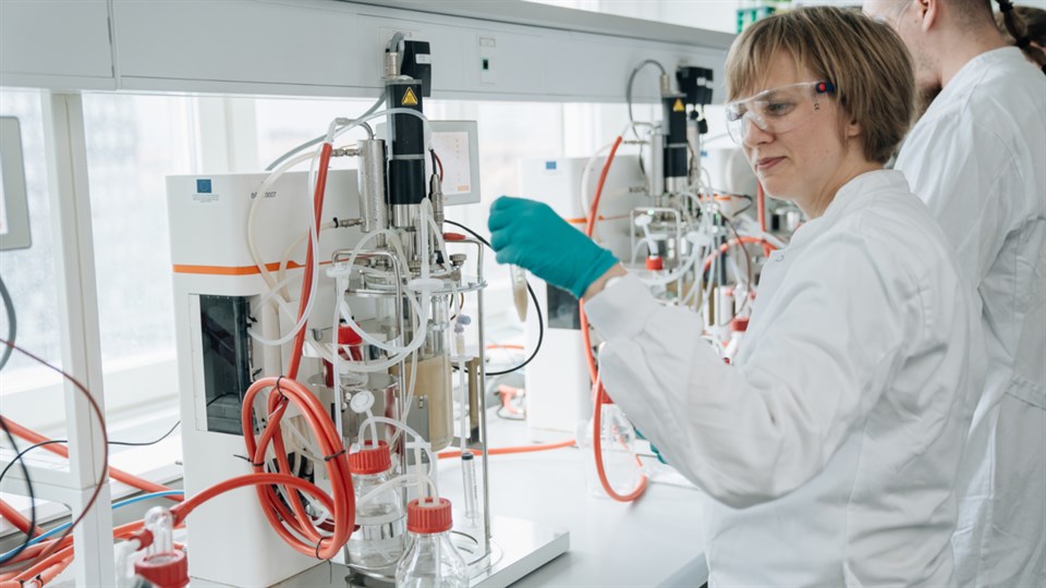 En kvinde i hvid kittel og blå gummihandsker arbejder ved en lille bioreaktor i et laboratorie