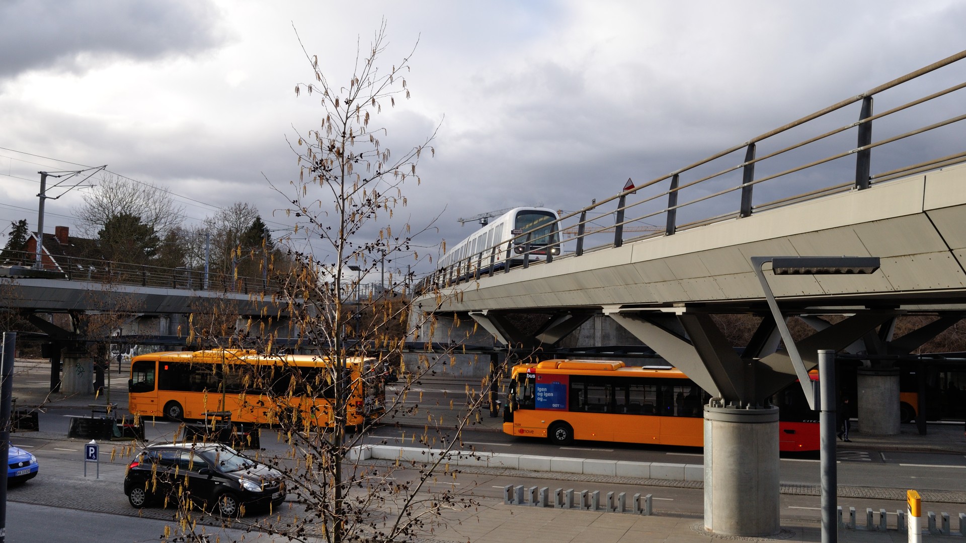 Foto af to Movia-busser på vejen ved Flintholm station. På broen over vejen kører et metrotog. 
