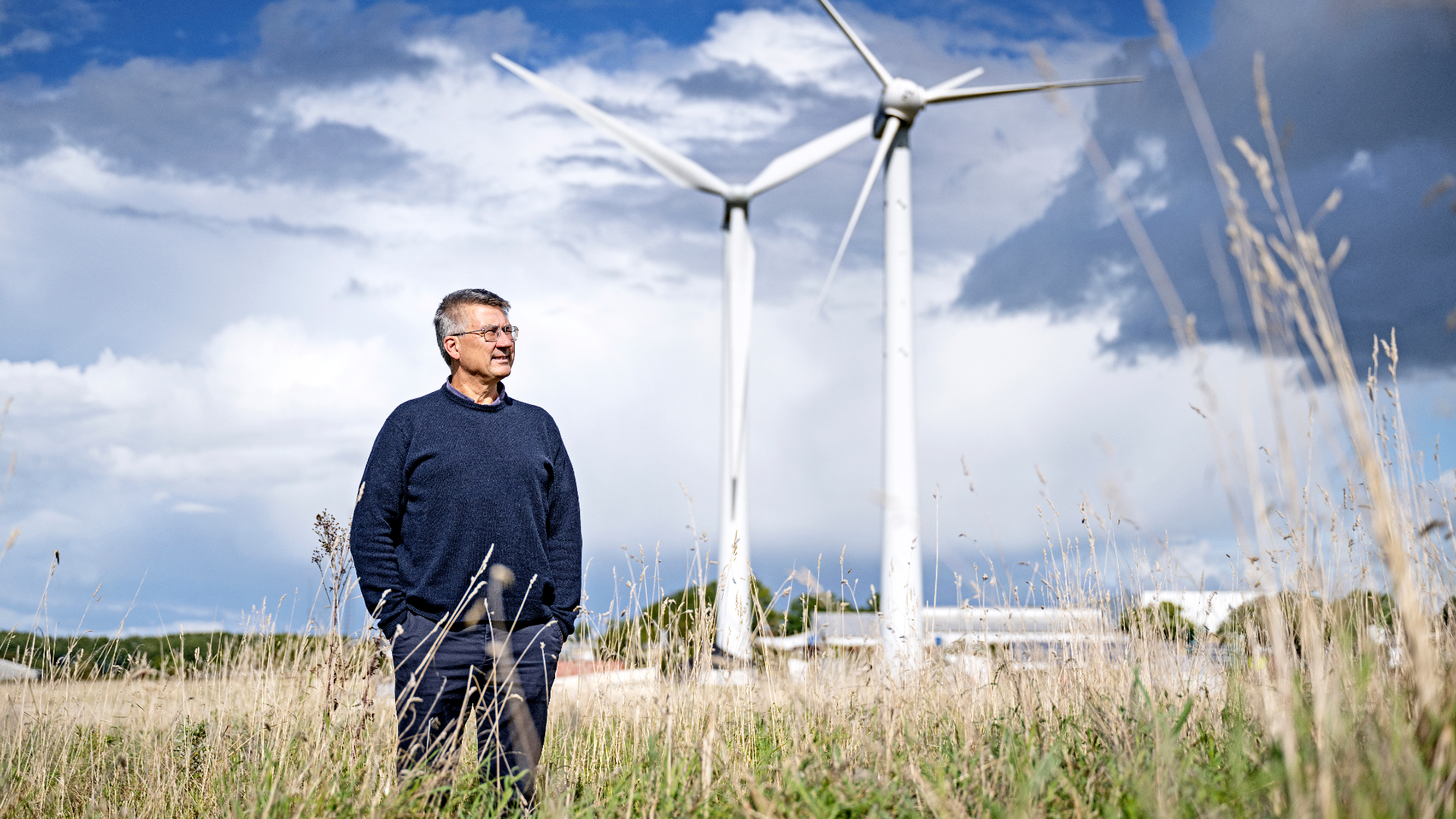 Portræt af Morten Jeppesen med to vindmøller i baggrunden og sensommergræs i forgrunden.