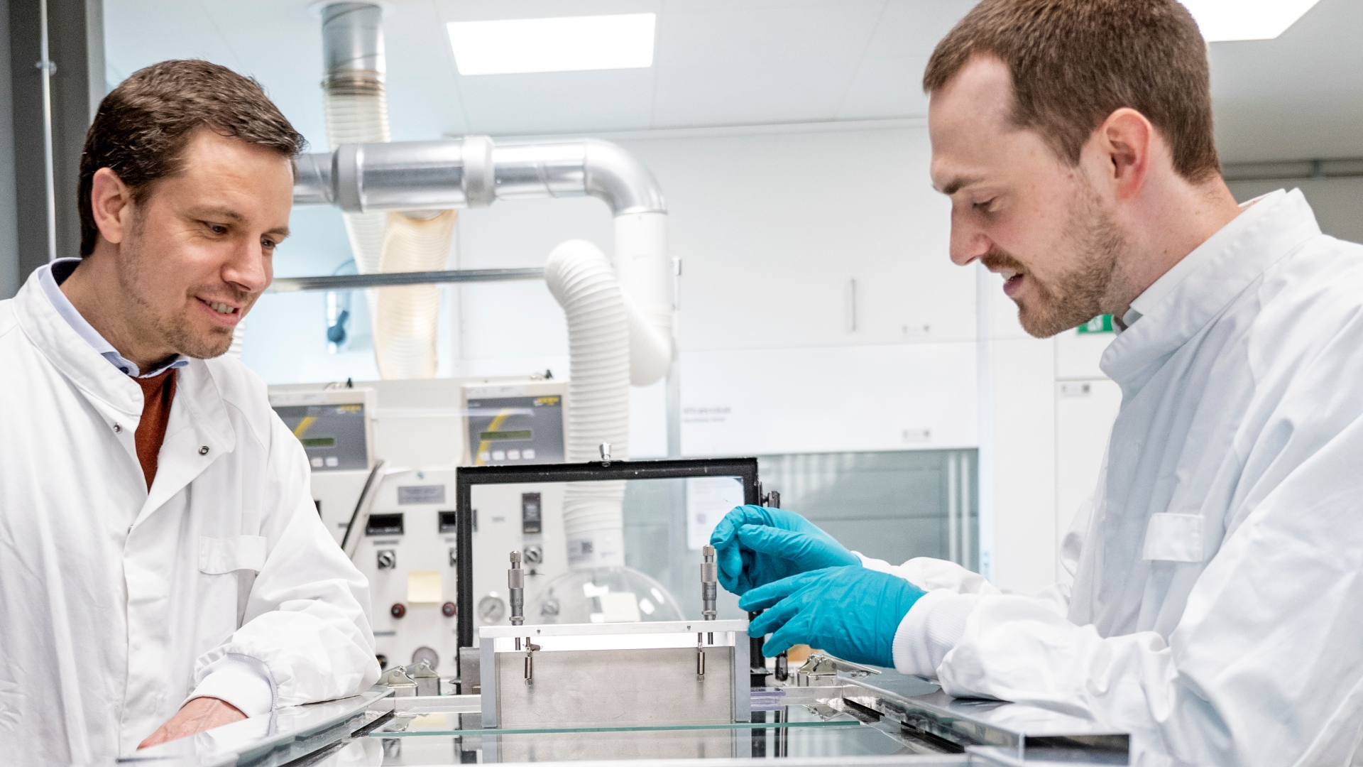 To mænd i hvide kitler i laboratorium. Den ene mand har blå handsker og rør ved et instrument i mellem dem, som de begge kigger på. 