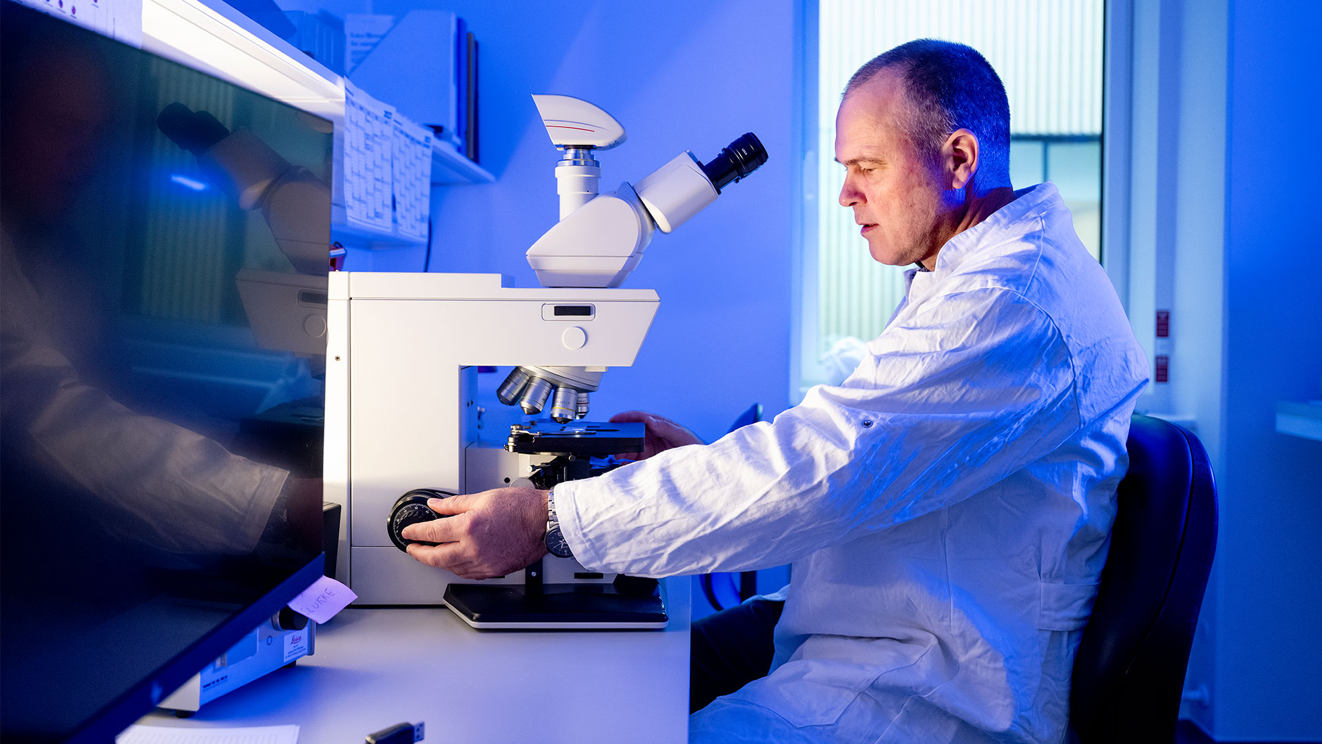 Man in a white lab coat is sitting at a microscope in a laboratory. Foto: Bax Lindhardt