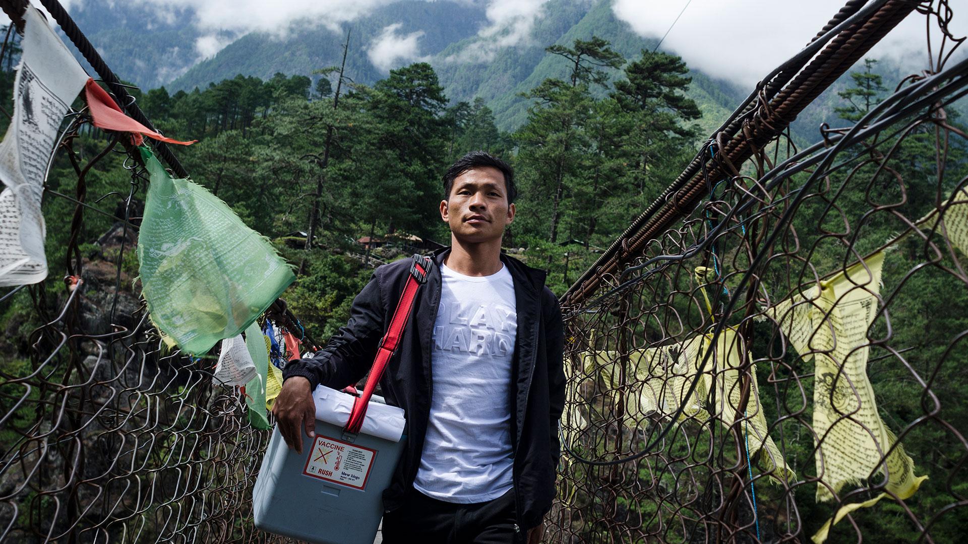 A man walks over a suspension bridge in a mountainous region with a chilly bin with vaccines across his shoulder.