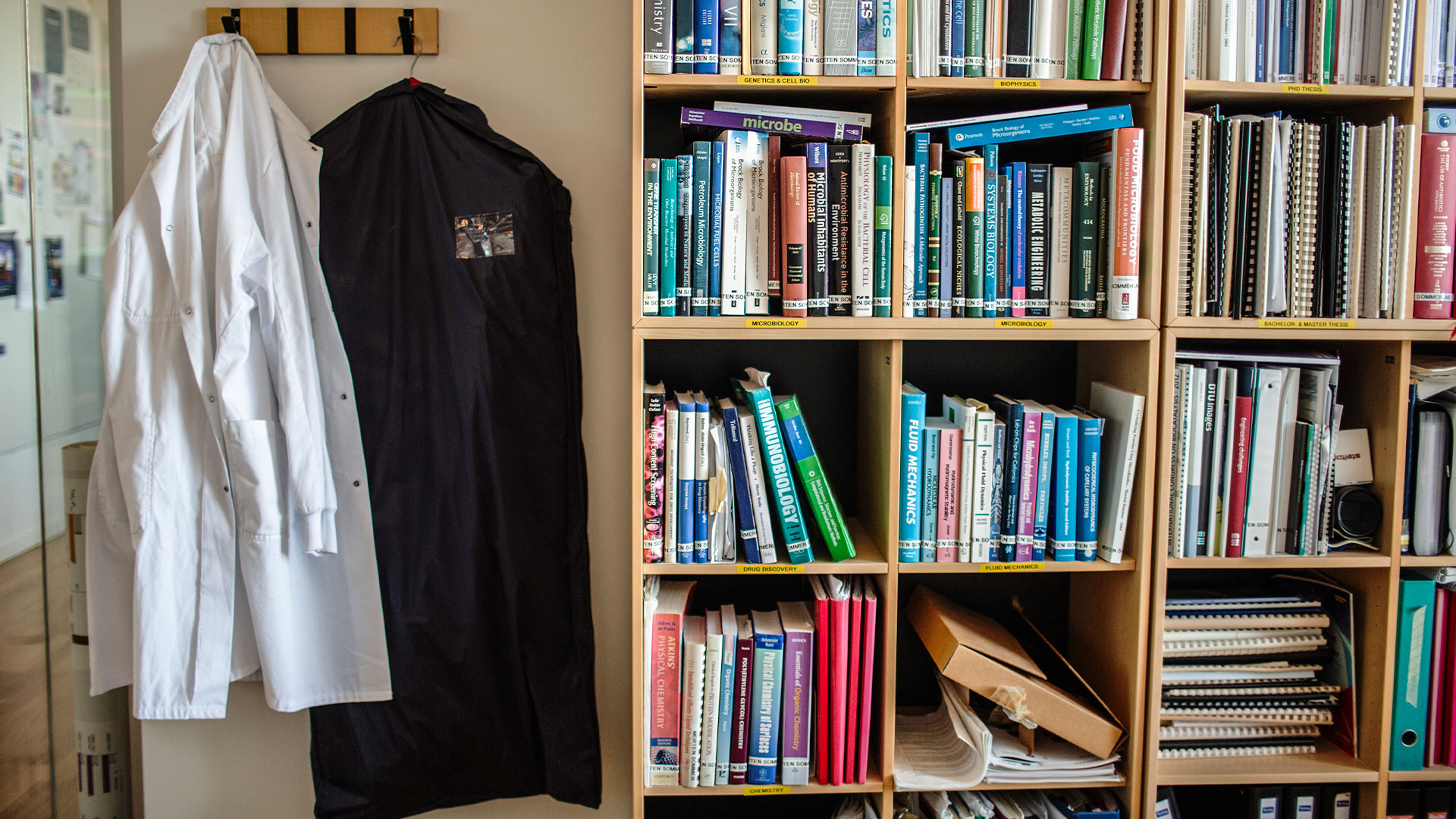 Room with bookshelves filled with books and a coatrack on which a lab coat and a suit bag is hanging. Photo: Ditte Valente