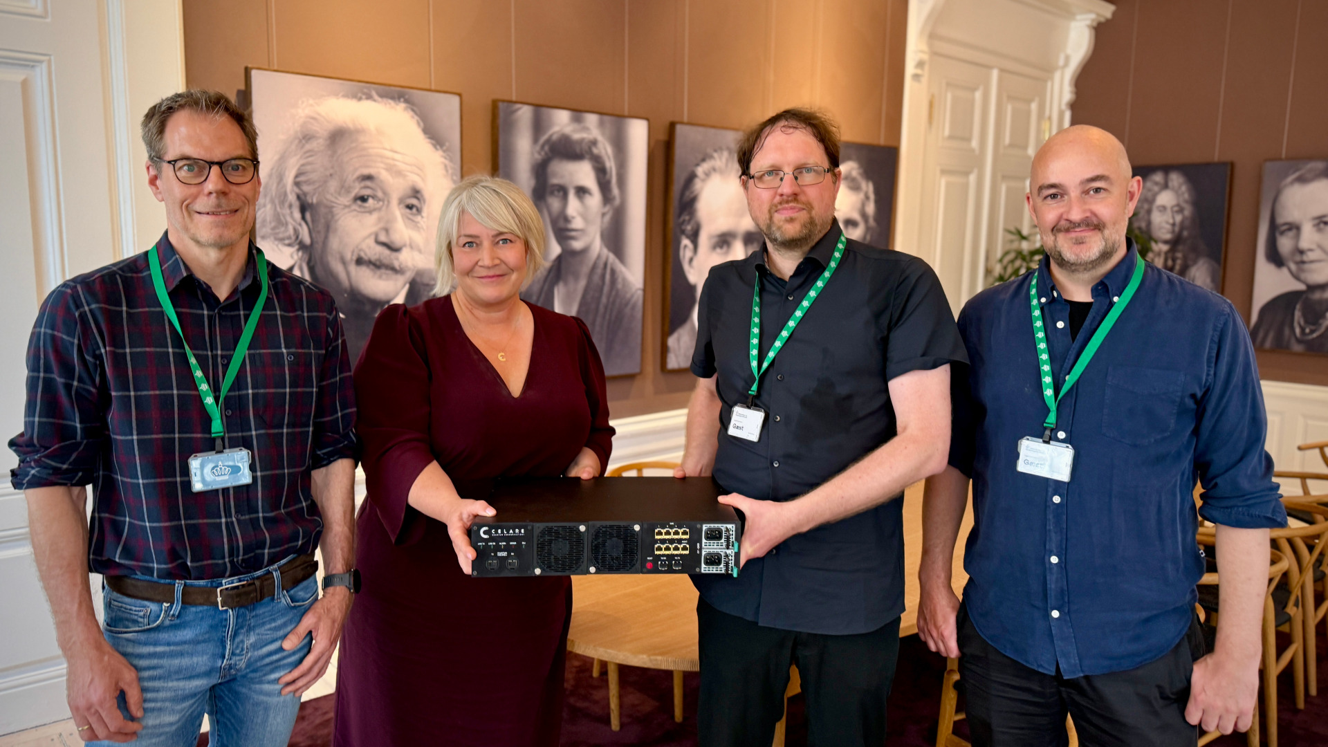 Four people standing on a row, 3 men and one woman. The woman and man in the middle hold a device used for producing quantum keys. It is a flat black box.